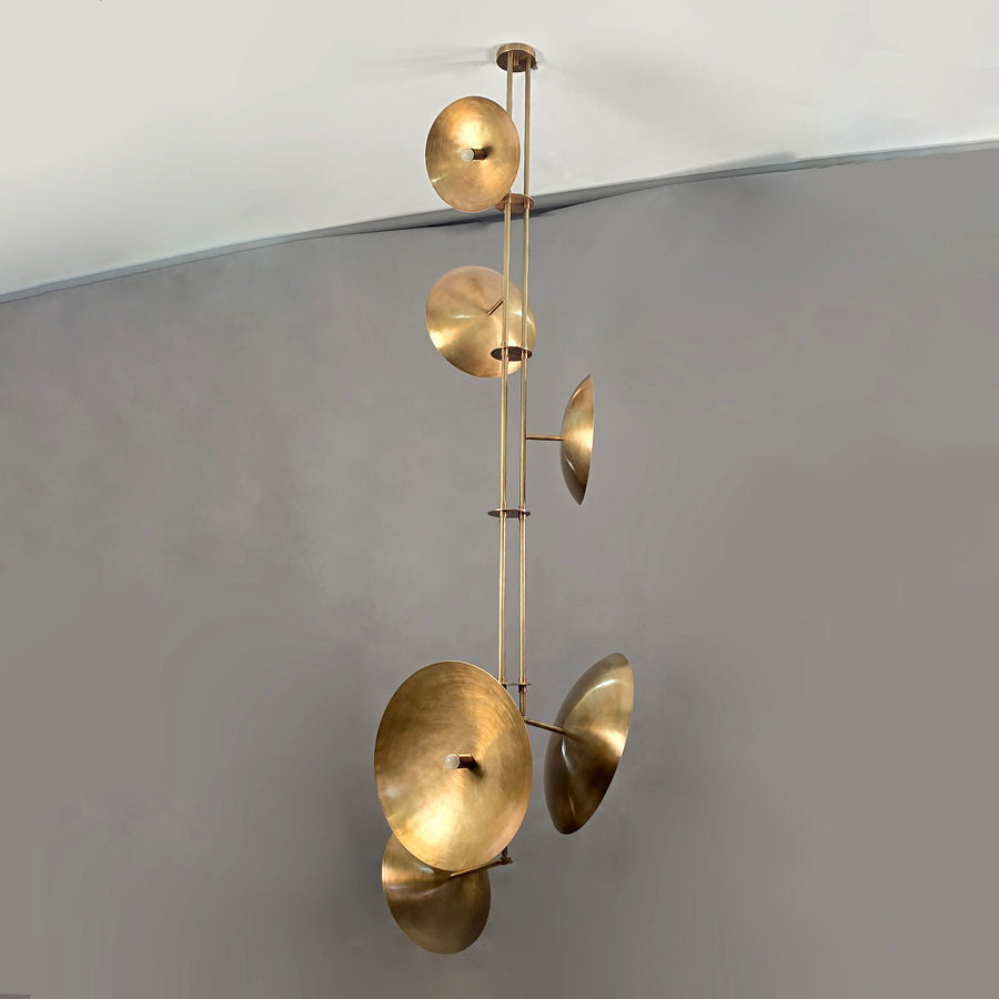 Vertical brass chandelier with six disc-shaped shades, showcasing sculptural form and warm ambient lighting.