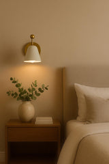 White shade brass wall sconce mounted above bedside table with vase and bed setup.
