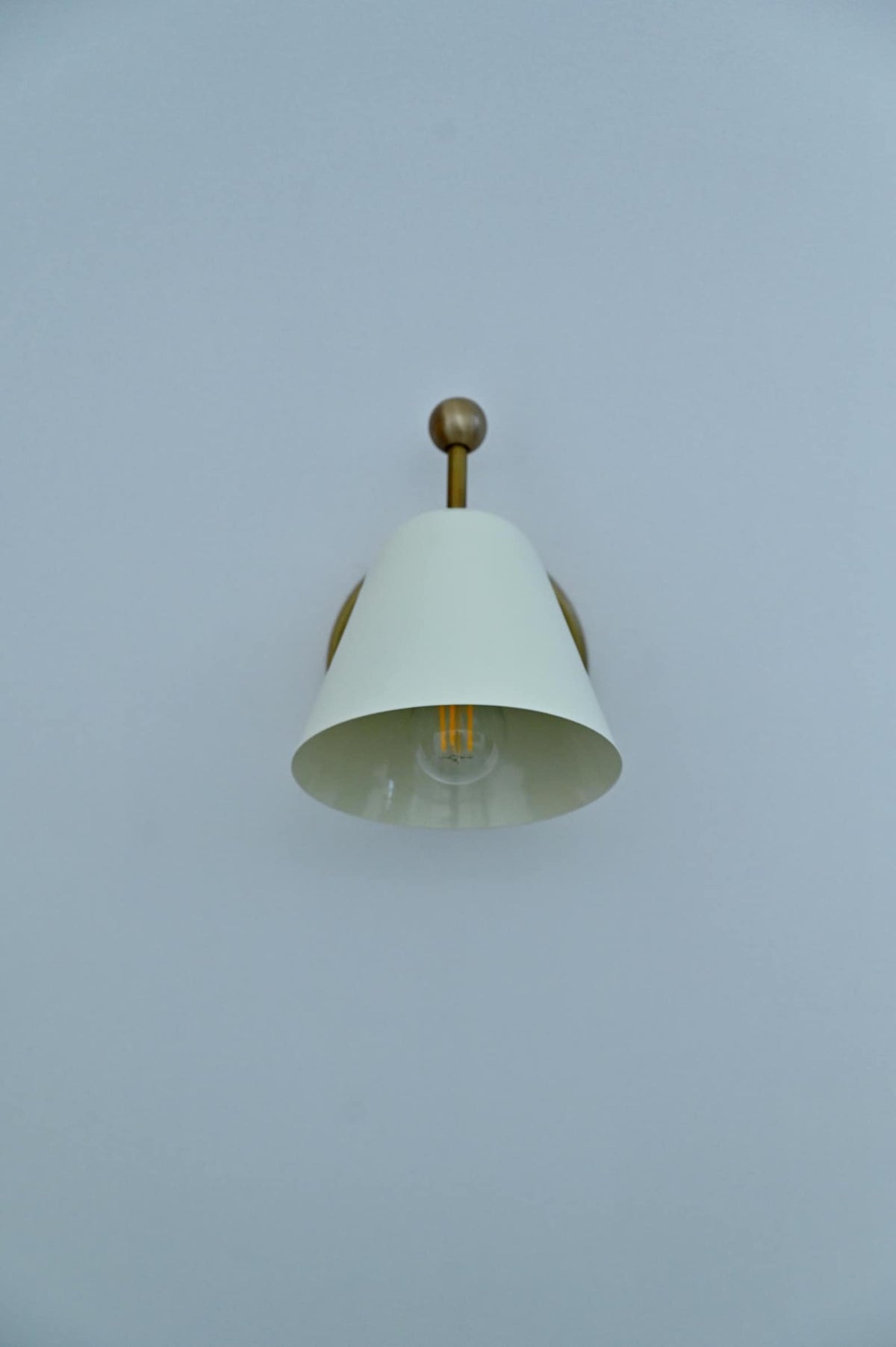 Front view of white conical shade brass wall sconce with exposed bulb inside.