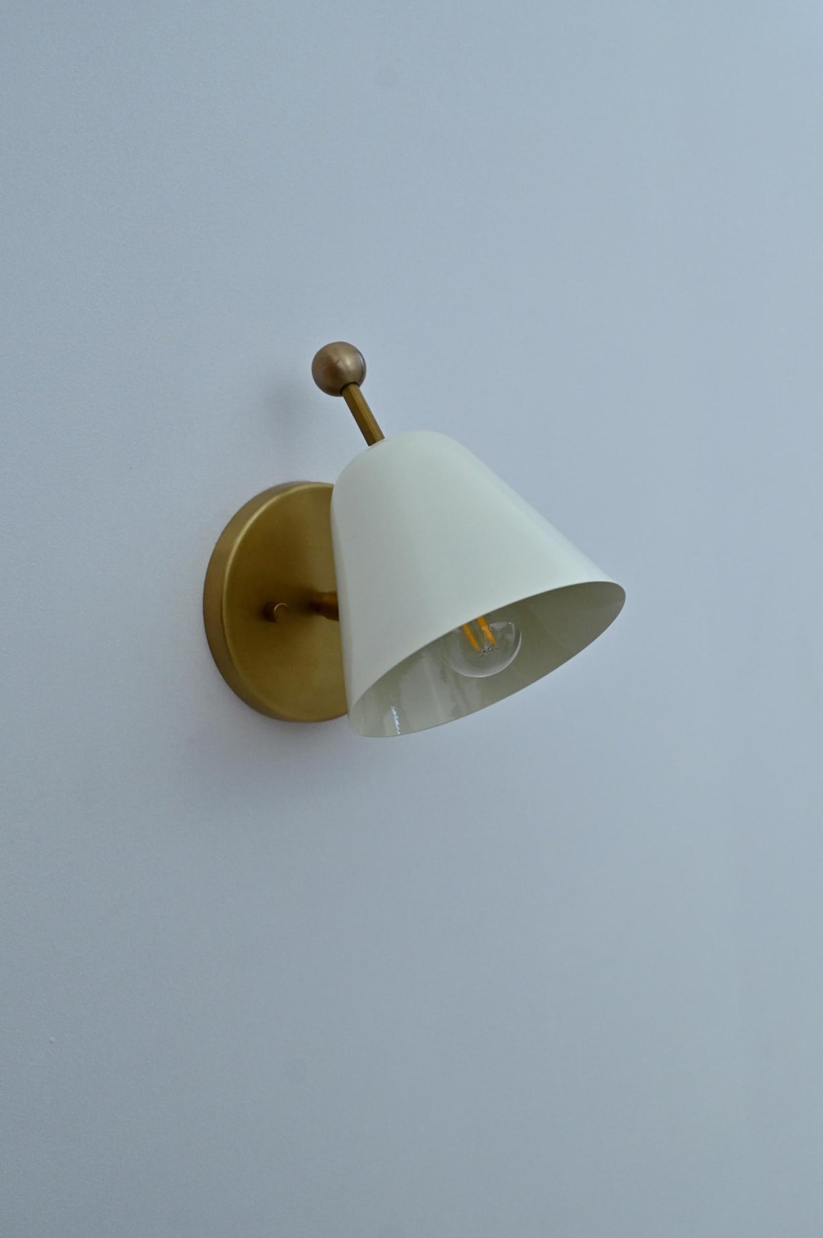 Side angled view of brass wall sconce with white conical shade on round brass base.
