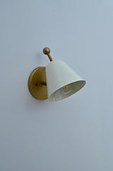 Side angled view of brass wall sconce with white conical shade on round brass base.
