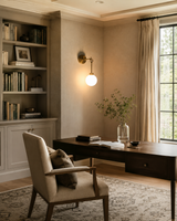 Windsor Modern Brass Globe Wall Sconce installed in a sophisticated home office, casting warm ambient light above a wooden desk with bookshelves, decorative accents, and a large window.