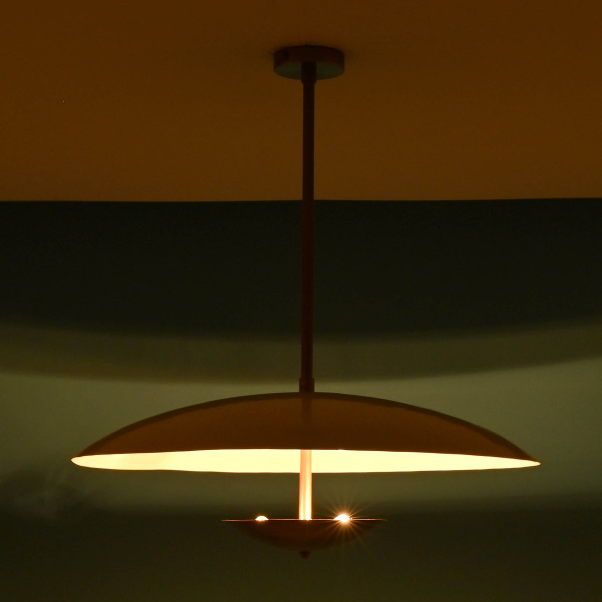 Ambient brass pendant light with wide dome shade glowing in a dark room, radiating sculpted modern lighting design.