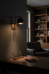 Black shade brass wall sconce installed beside a bed with warm downward reading light and brass patina finish