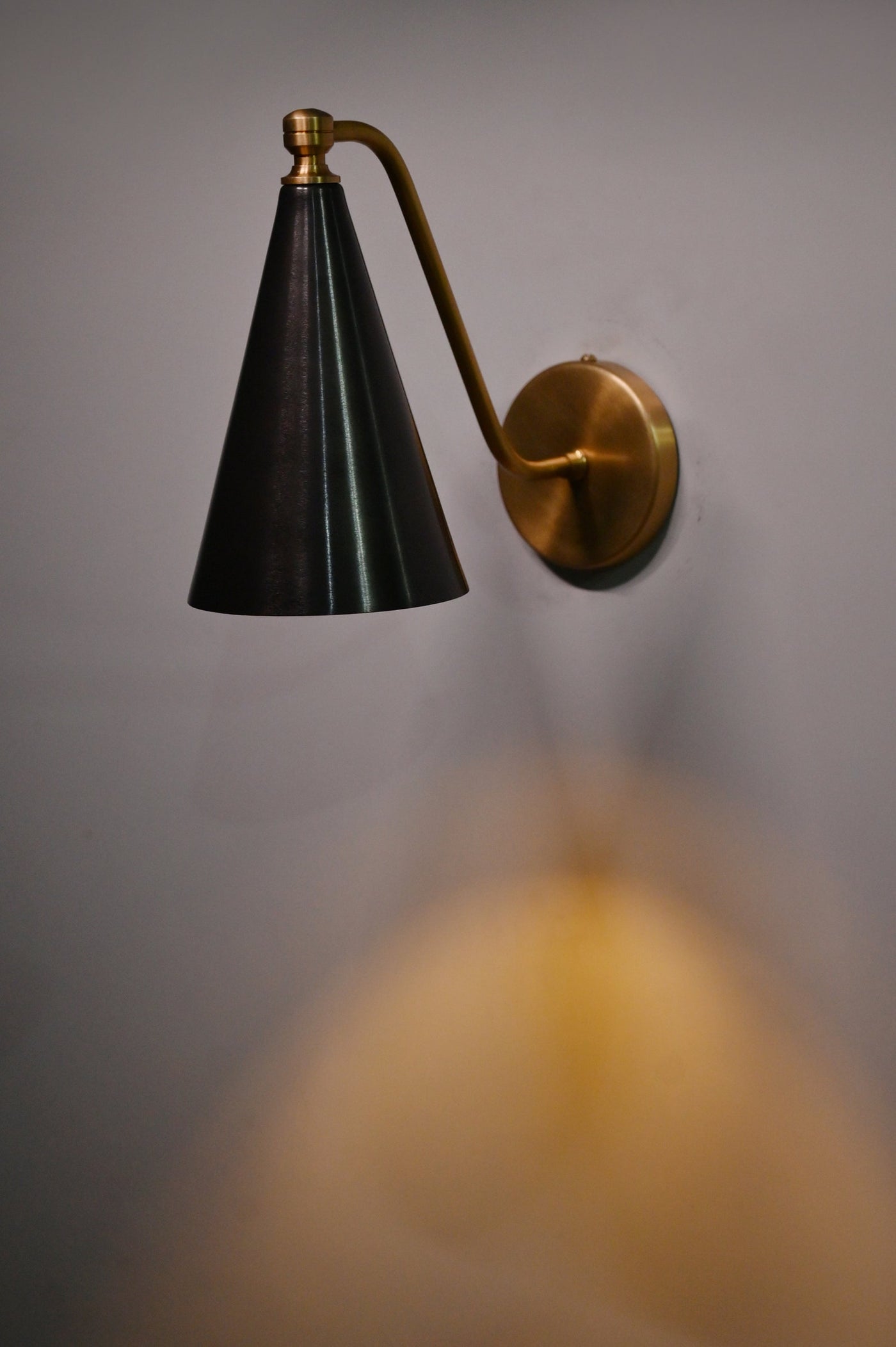 Blackened brass cone wall light casting a warm ambient glow, featuring brushed brass arm and round wall mount.