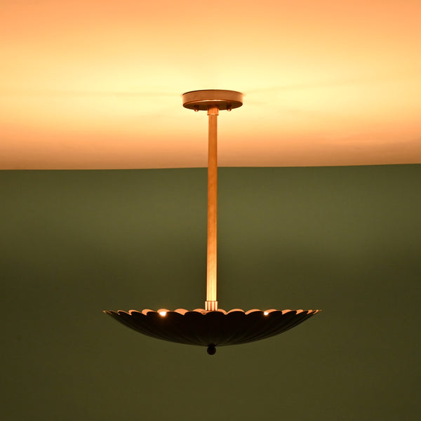 Sculptural floral pendant light casting a warm glow on the ceiling with raw brass rod and blackened brass shade.