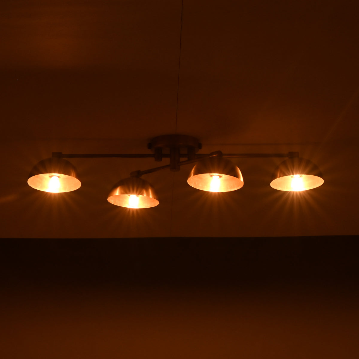Low-profile handmade brass chandelier with four illuminated dome shades, casting golden lighting on the ceiling.