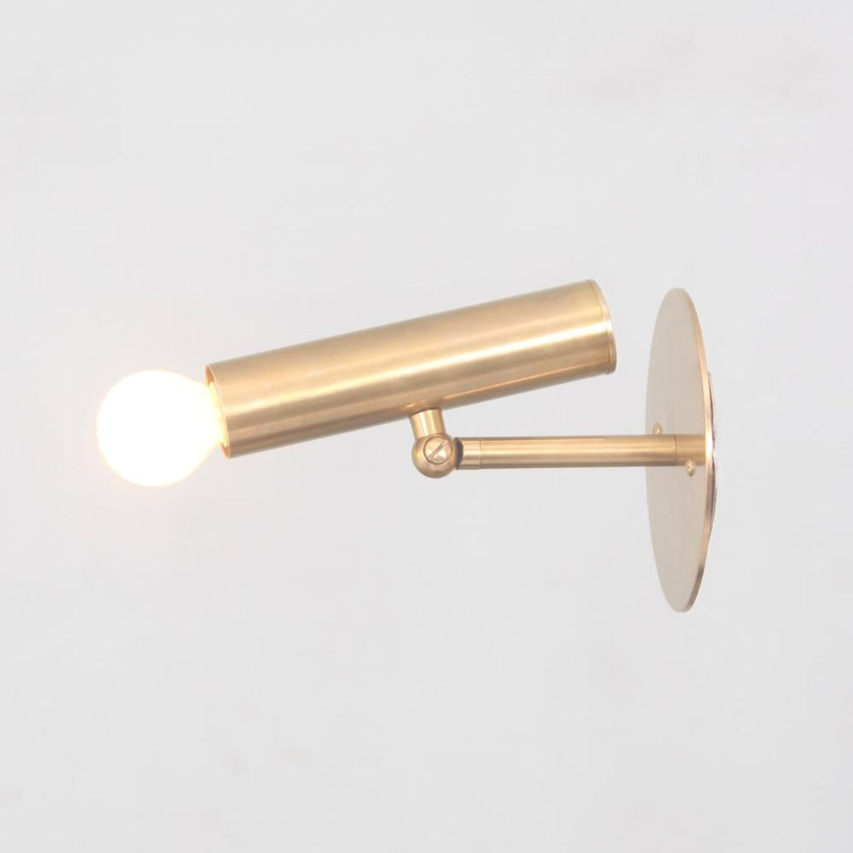 Brass wall spotlight with extended cylindrical body and glowing bulb, mounted on brass base