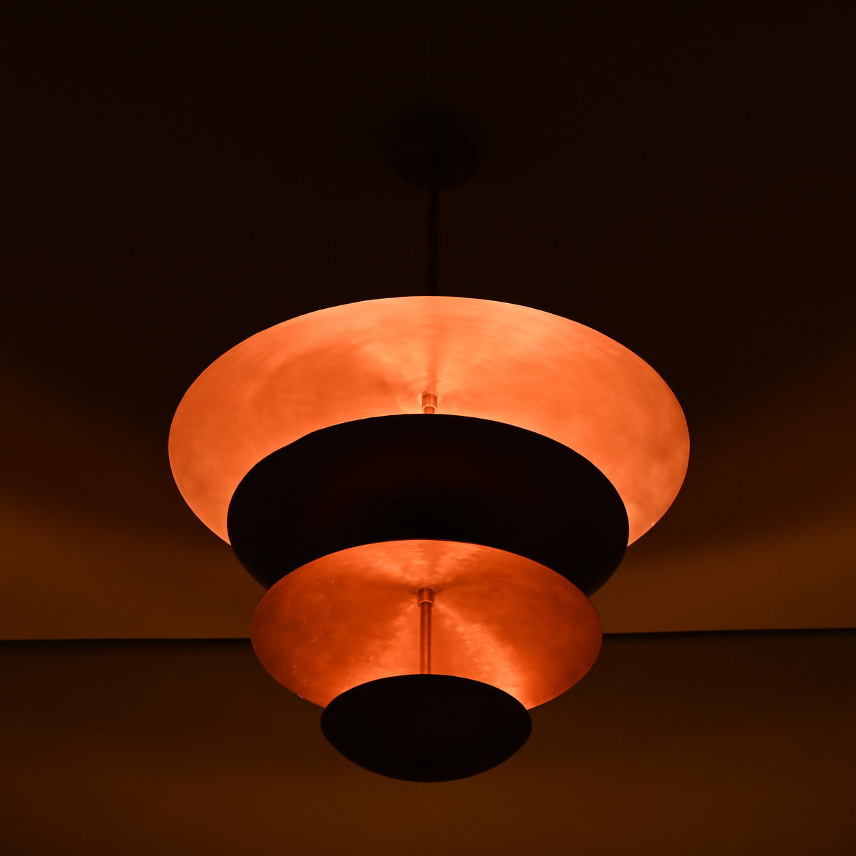 Night-shot of the brass bowl chandelier illuminated with warm indirect lighting across its stacked copper shades.