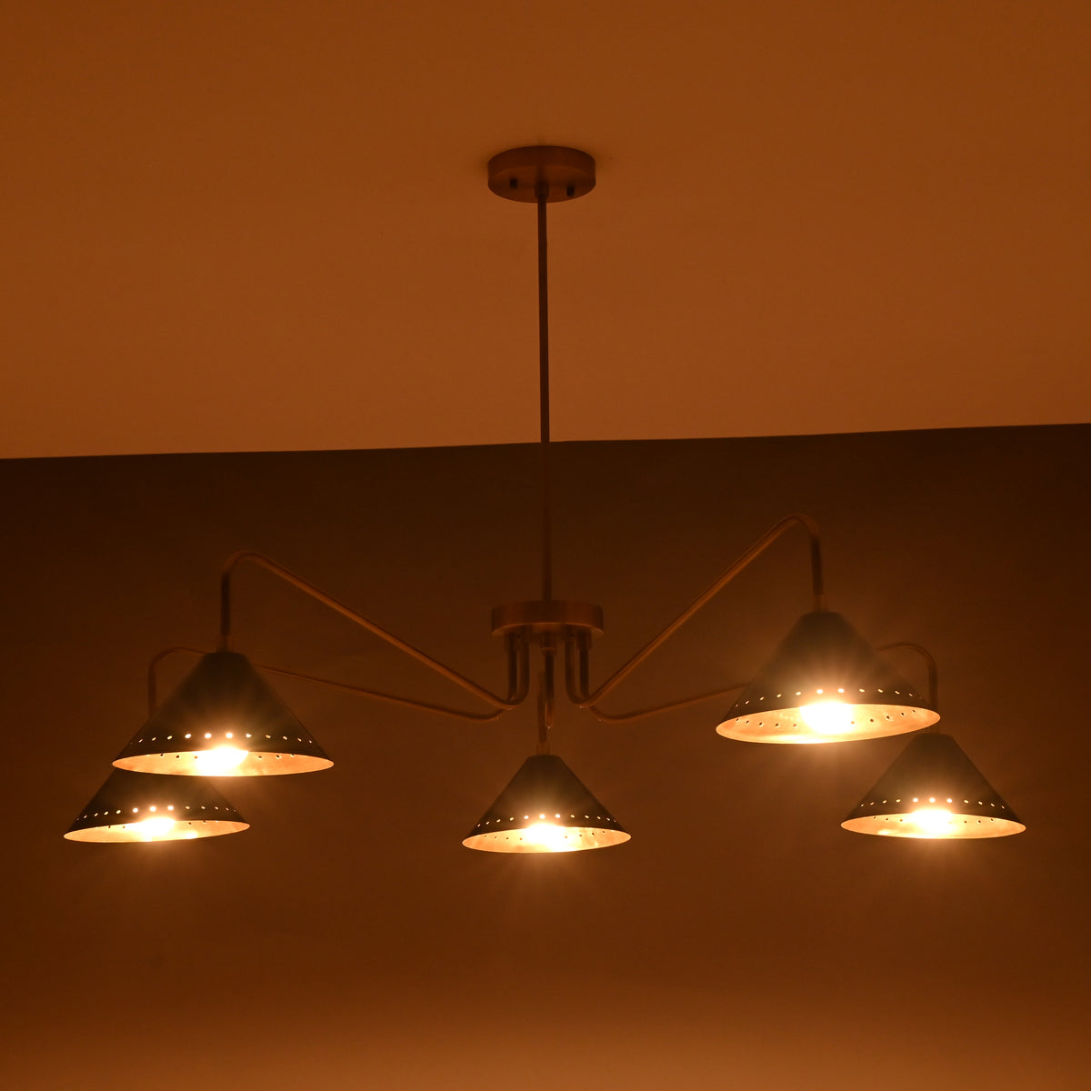 Evening ambience view of brass cone chandelier with five perforated black shades casting warm diffused light.