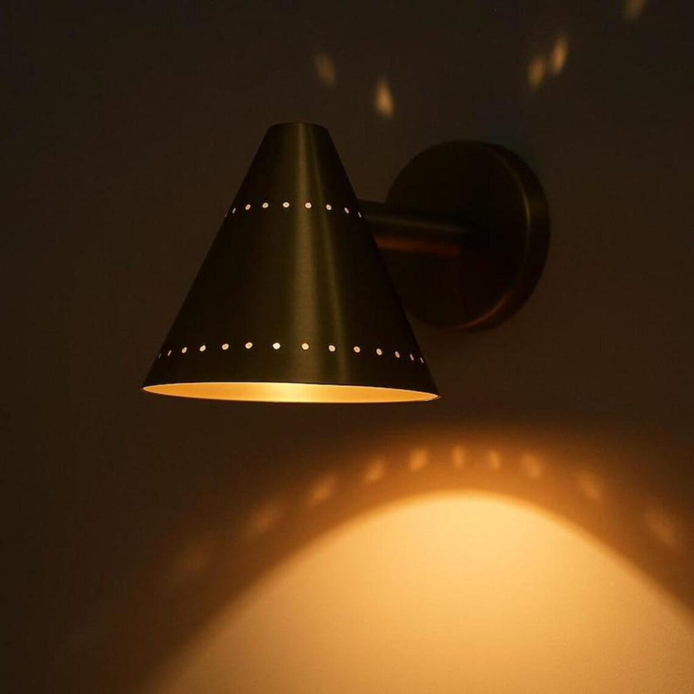 Minimalist brass cone wall sconce casting warm ambient light, highlighting perforated detailing and soft indirect illumination.