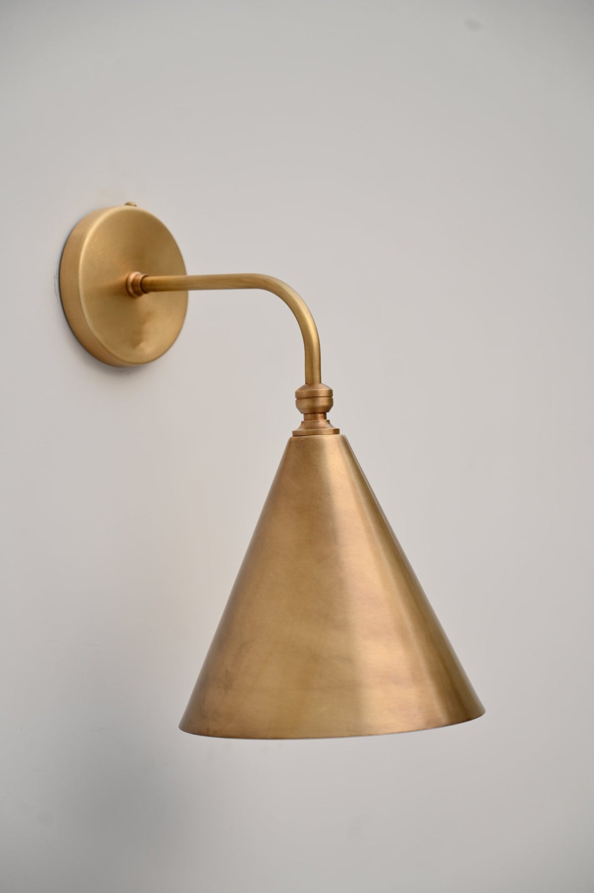 Angled view of brass cone wall sconce with curved arm and circular backplate, handcrafted by Brass Leaf Studio