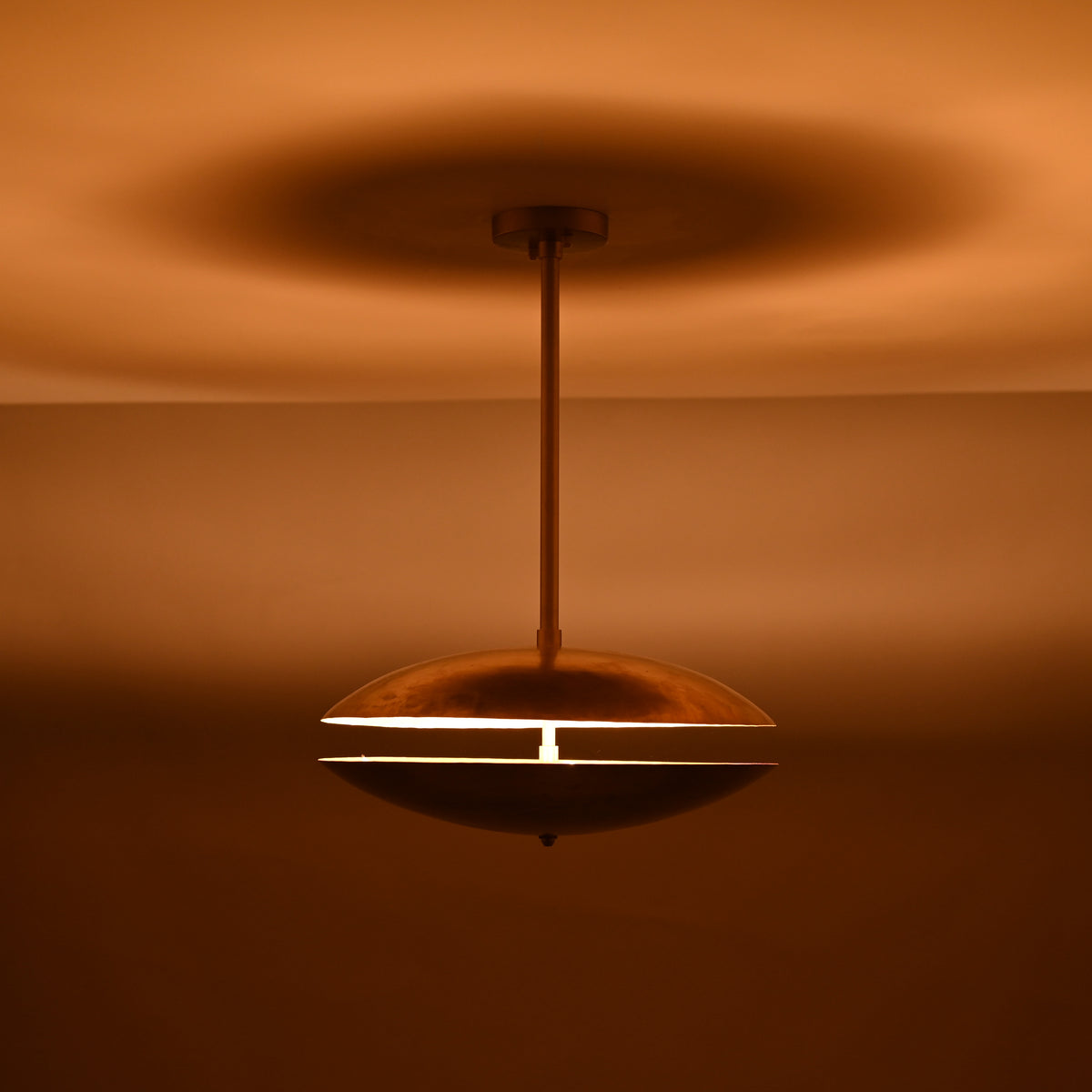 Brass disc pendant light with narrow slit illumination glowing in a warm amber tone, showcasing dramatic ambient lighting in the dark.