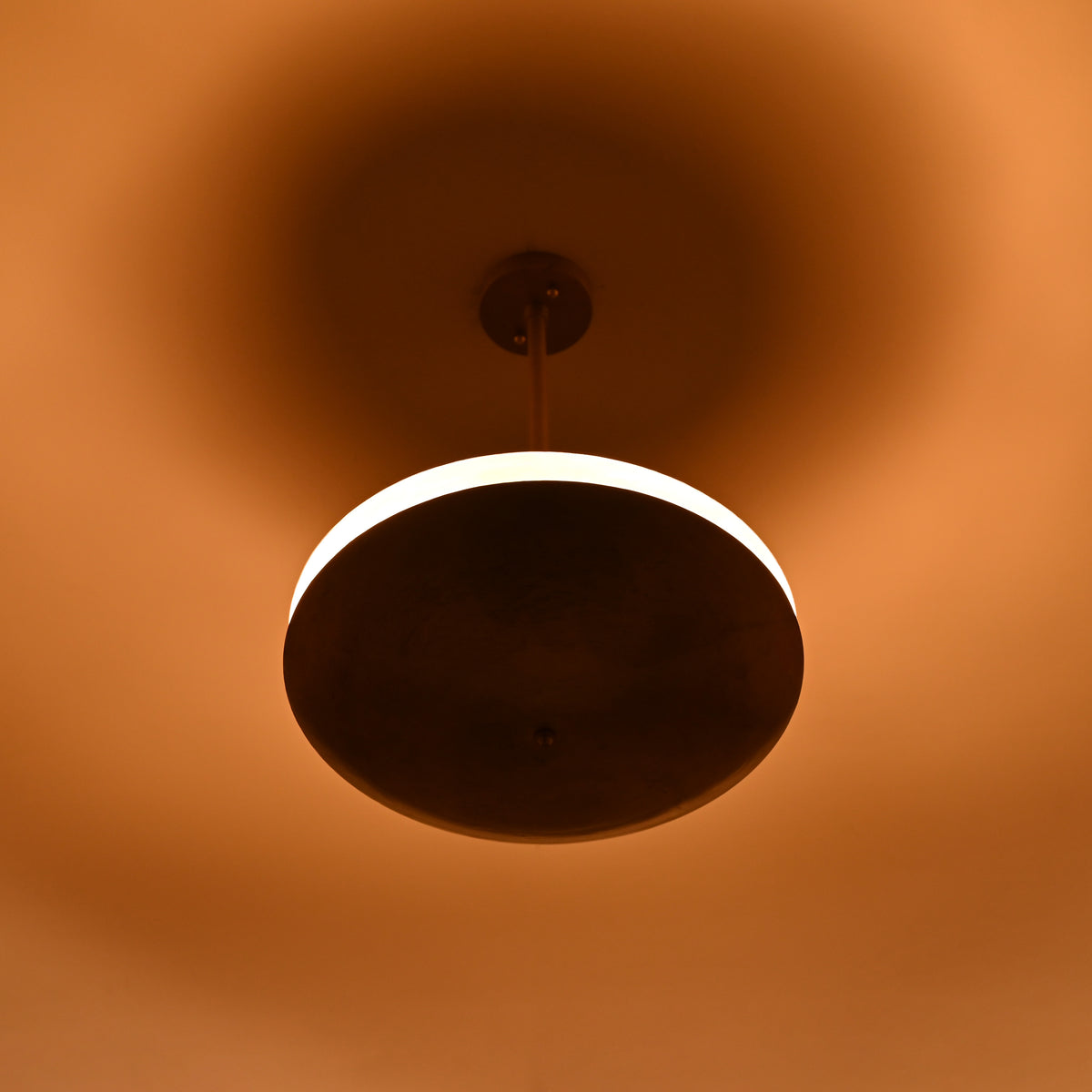 Brass disc pendant light photographed in low light, showing bright illuminated ring and soft ambient glow on ceiling and surroundings.