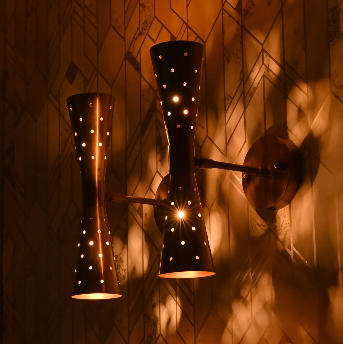 Close-up of two brass double cone wall sconces lit, casting warm patterned light on wall