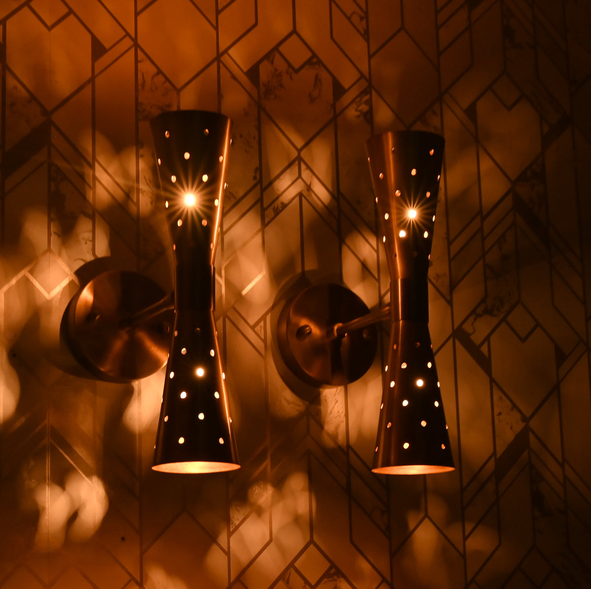 Two brass double cone wall sconces glowing warmly, creating decorative light patterns on wall