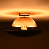 Exposed-bulb view of polished and blackened brass flushmount light, glowing warmly and casting soft diffused shadows on the ceiling.