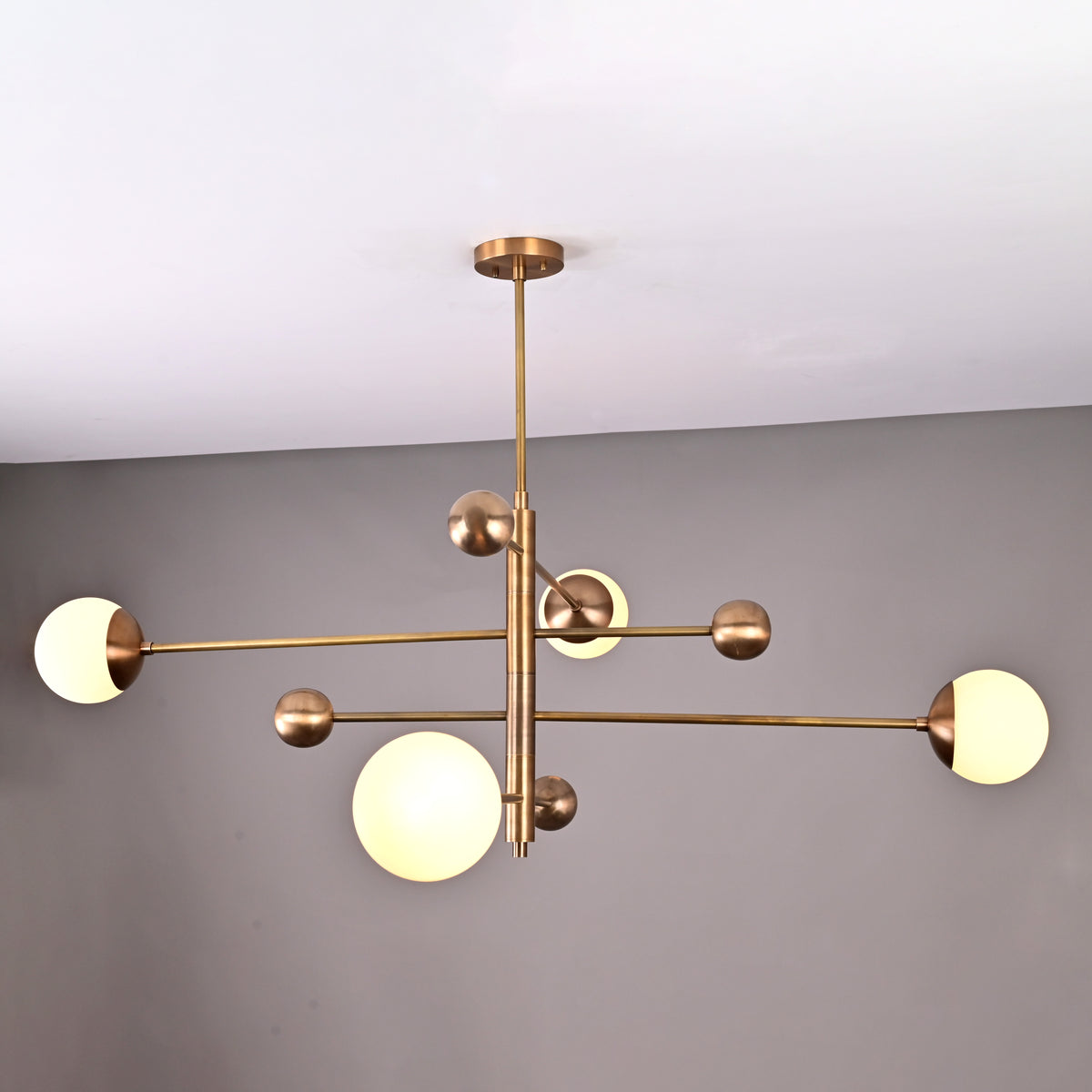 Angled view of modern brushed brass chandelier featuring four opal glass globes on minimalist linear arms.