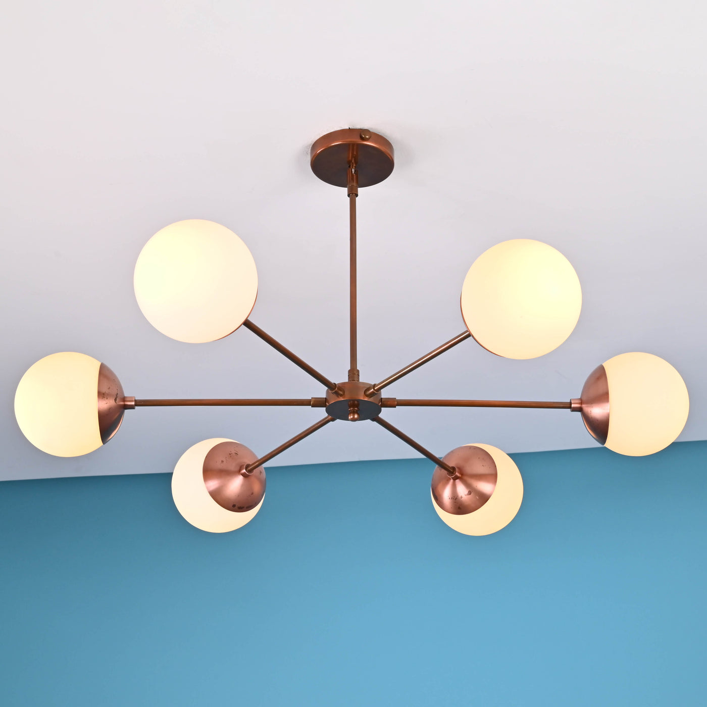 Six-light modern brass chandelier with spherical frosted glass globes and copper finish arms mounted on ceiling against blue wall.
