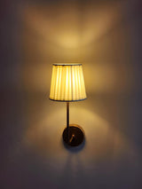 Handmade brass wall sconce by Brass Leaf Studio with pleated fabric shade glowing warmly against a wall.