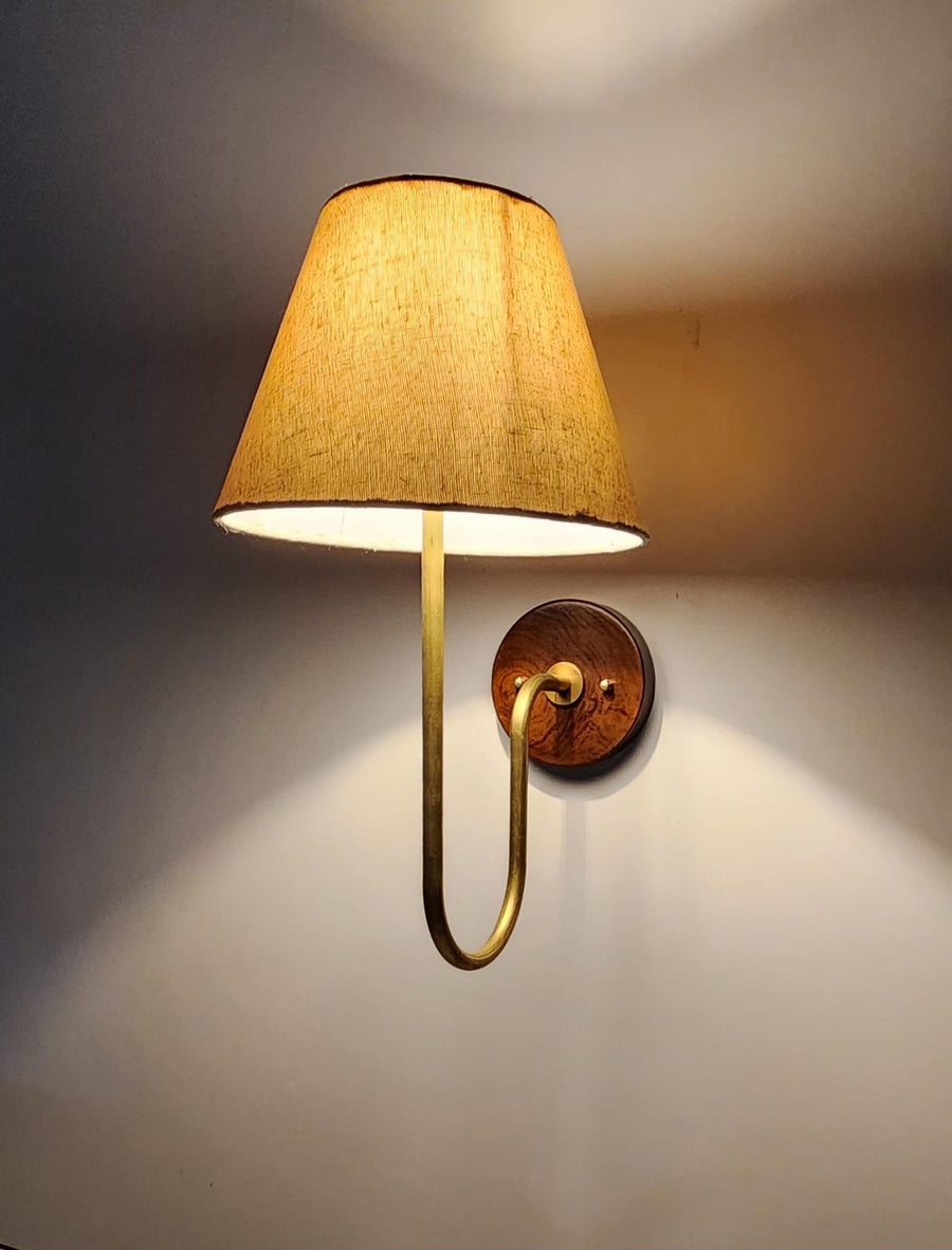 Handmade brass wall light with curved arm and beige fabric shade, emitting warm ambient light against wall.