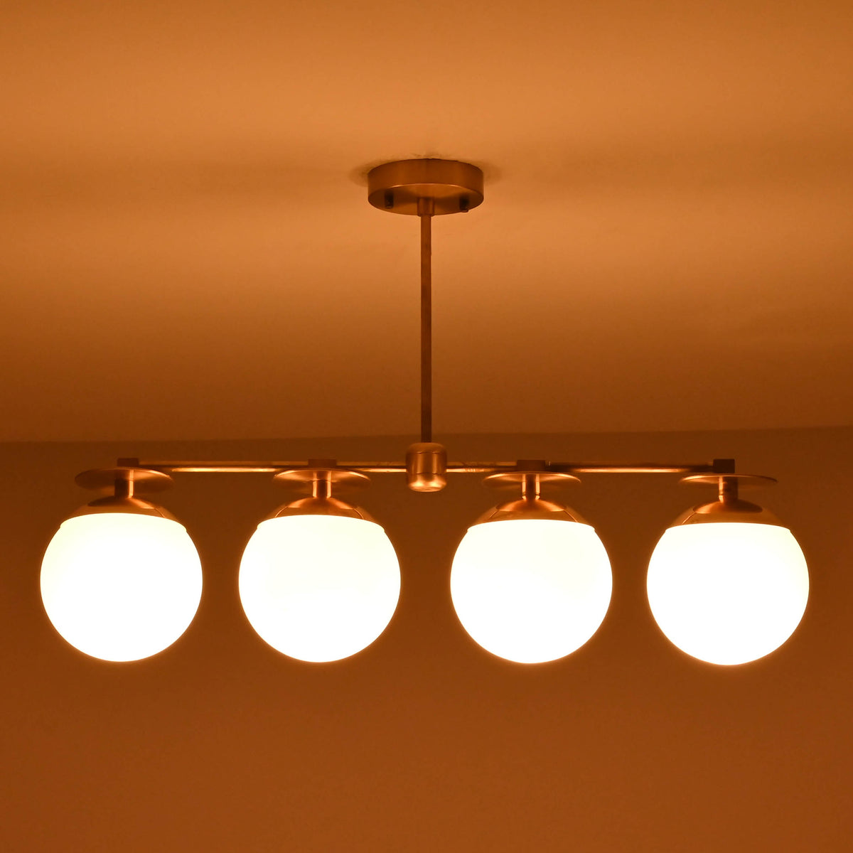 Front-facing view of brass linear chandelier with four opal glass globes in a low-light interior setting.
