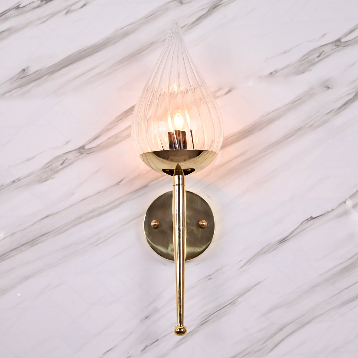 Polished brass wall sconce with clear teardrop glass shade installed on white veined marble surface