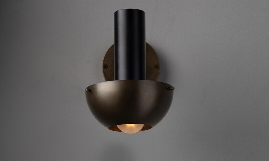 Modern brass wall lamp with sculptural bowl shade and cylindrical form, casting warm ambient light in a refined industrial design.