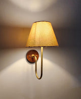 Handmade brass wall light with curved arm and beige fabric shade, emitting warm ambient light against wall.