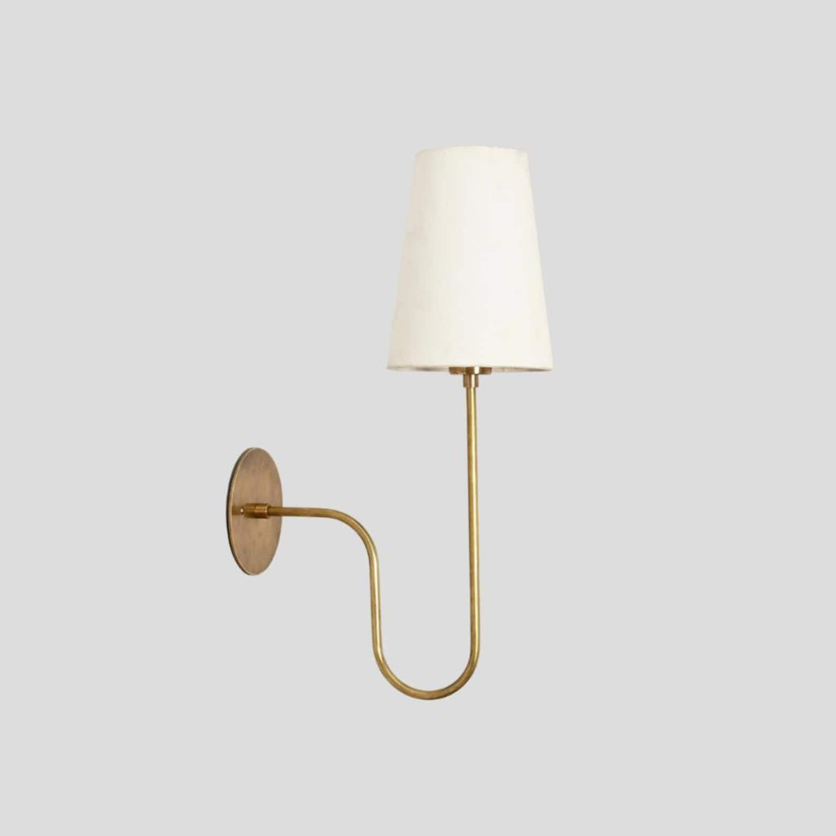 Minimalist brass wall light featuring a round backplate, curved brass arm, and soft fabric shade for warm ambient lighting.