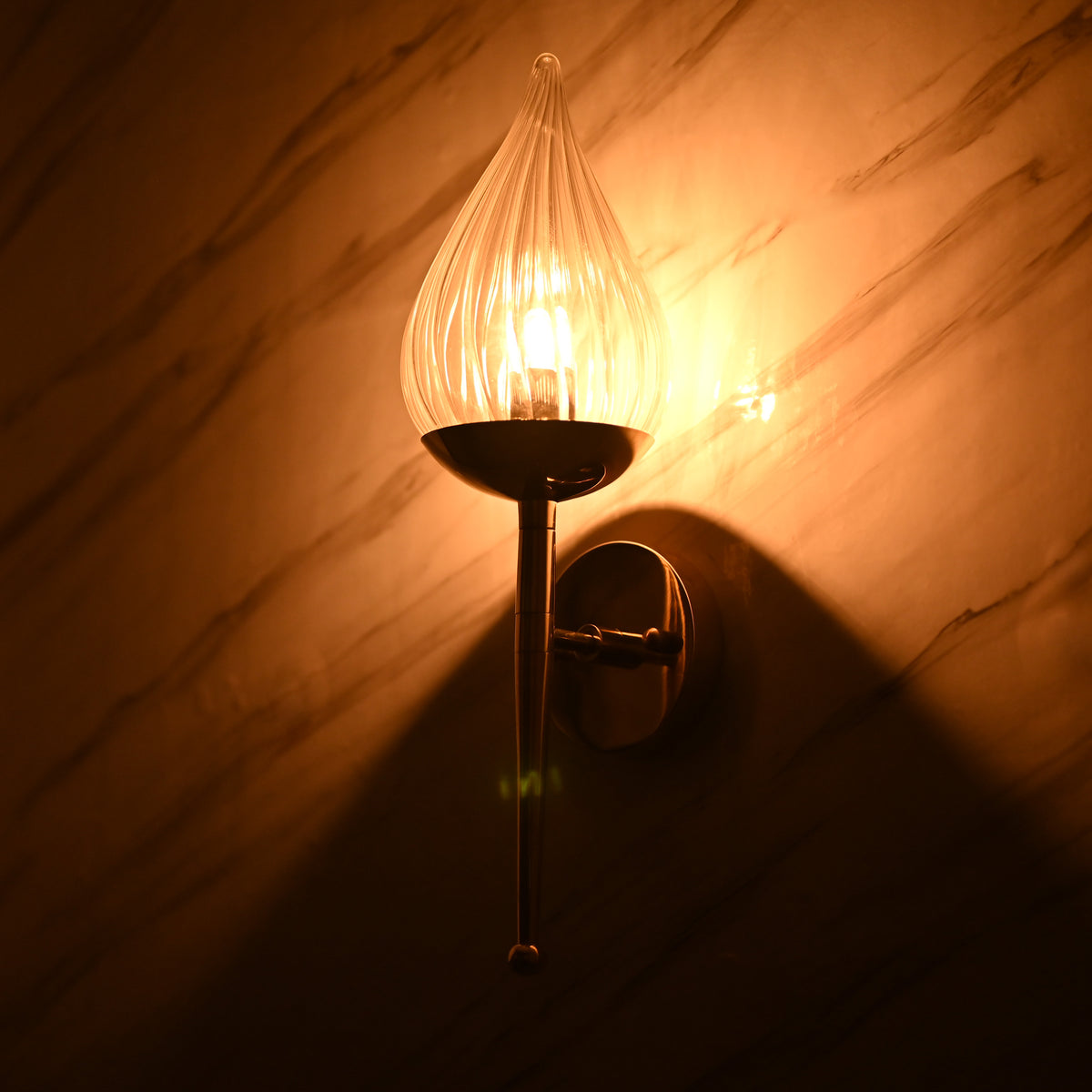 Warm glowing light from fluted glass teardrop sconce on brass base casting ambient shadows on marble wall