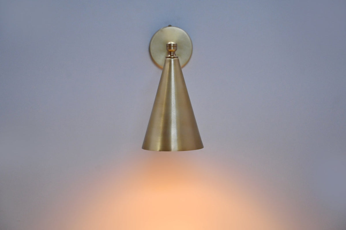 Front view of brass wall sconce with cone shade casting warm downward light, perfect for modern minimalist interiors.