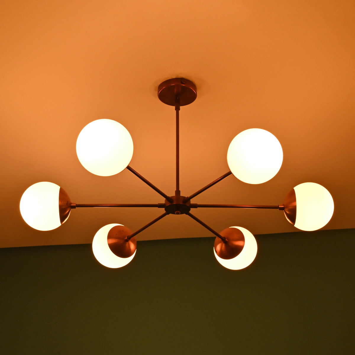 Modern brass chandelier with six frosted glass globes glowing warmly on ceiling in a dimly lit room.