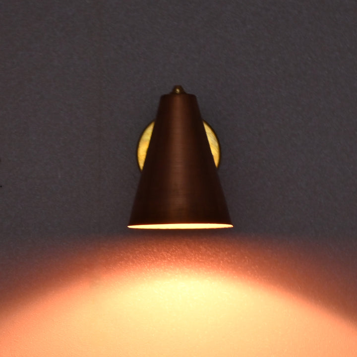 Brass and copper wall sconce lit in a dark setting, casting a warm ambient glow on the wall.