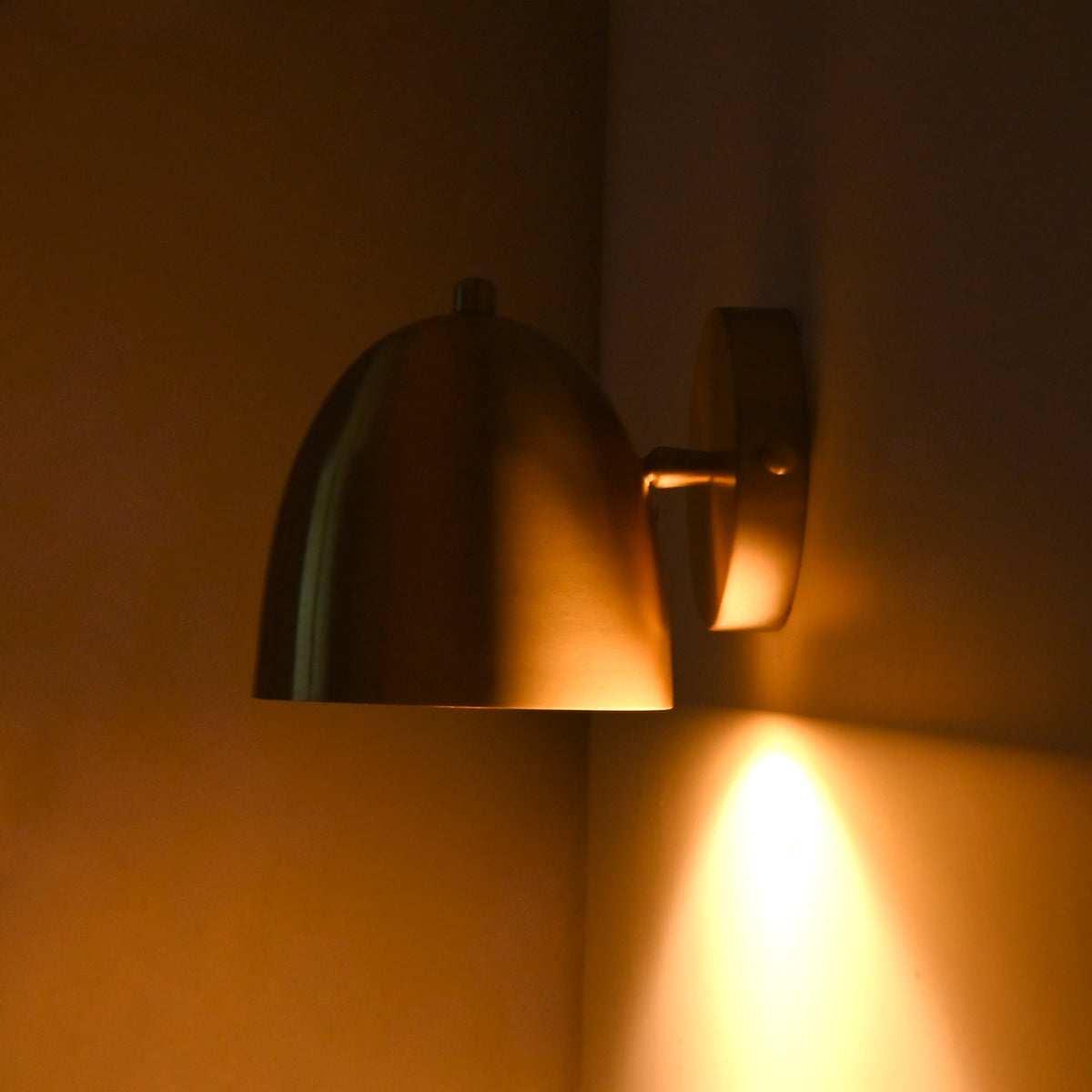 A modern dome-shaped wall sconce with a brushed brass finish, mounted on a dark wall and casting a warm golden light downward.