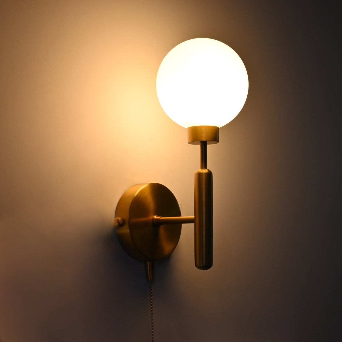 Brass wall sconce with a glowing round glass globe shade and pull chain, mounted on a wall and illuminated.
