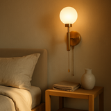 Brass pull chain globe wall sconce glowing warmly above a bedside table with folded throw and ceramic vase, next to a bed with linen pillows.