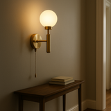 Raw brass pull chain globe wall sconce illuminated above a wooden console table with stacked books, in a hallway setting.