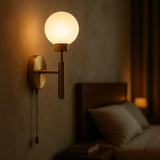 Raw brass pull chain globe wall sconce illuminated beside a bed, casting warm light near a dark wood headboard and nightstand.