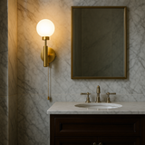 Brass pull chain globe wall sconce illuminated beside a mirror in a marble-tiled bathroom, mounted above a dark wood vanity.