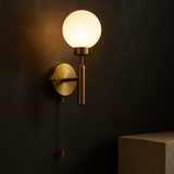 Raw brass pull chain globe wall sconce illuminated on a dark textured wall, with a frosted glass globe shade above a stone pedestal.