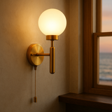 Illuminated raw brass pull chain globe wall sconce mounted on a dark textured wall, glowing softly with warm light.