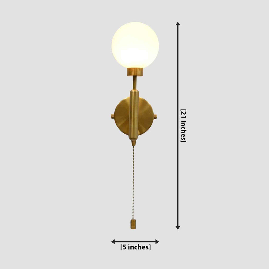Brass pull chain globe wall sconce illuminated beside a bed, casting warm light near a window with sheer curtains and a wooden nightstand.