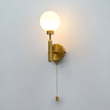 Raw brass wall sconce with round frosted glass globe shade, illuminated, and hanging pull chain switch against a white wall.