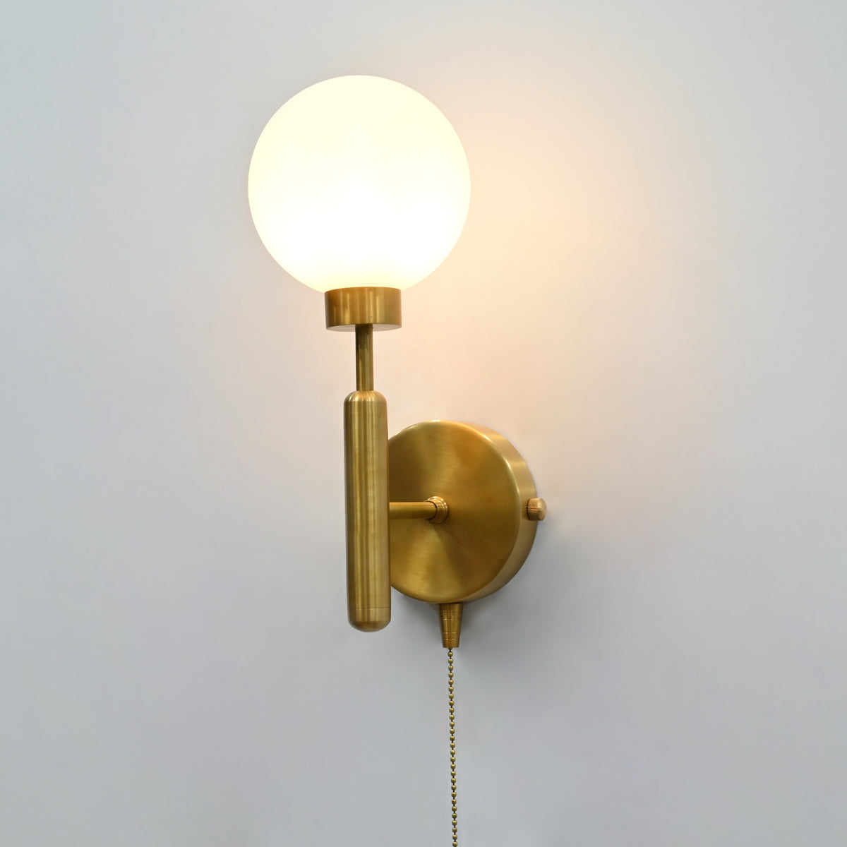 Raw brass wall sconce with a round frosted glass globe shade, illuminated, featuring a pull chain switch against a light gray wall.
