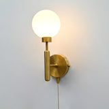 Raw brass wall sconce with a round frosted glass globe shade, illuminated, featuring a pull chain switch against a light gray wall.