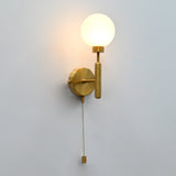 Raw brass wall sconce with round frosted glass globe shade, illuminated, and hanging pull chain switch, shown against a plain wall.