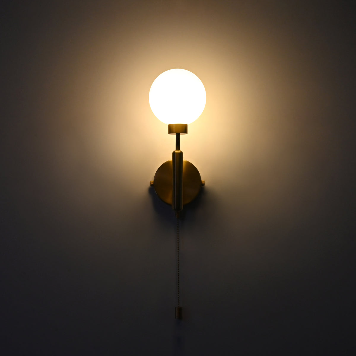 Pull chain globe wall sconce in raw brass finish, illuminated with a warm glow from the frosted glass globe shade.