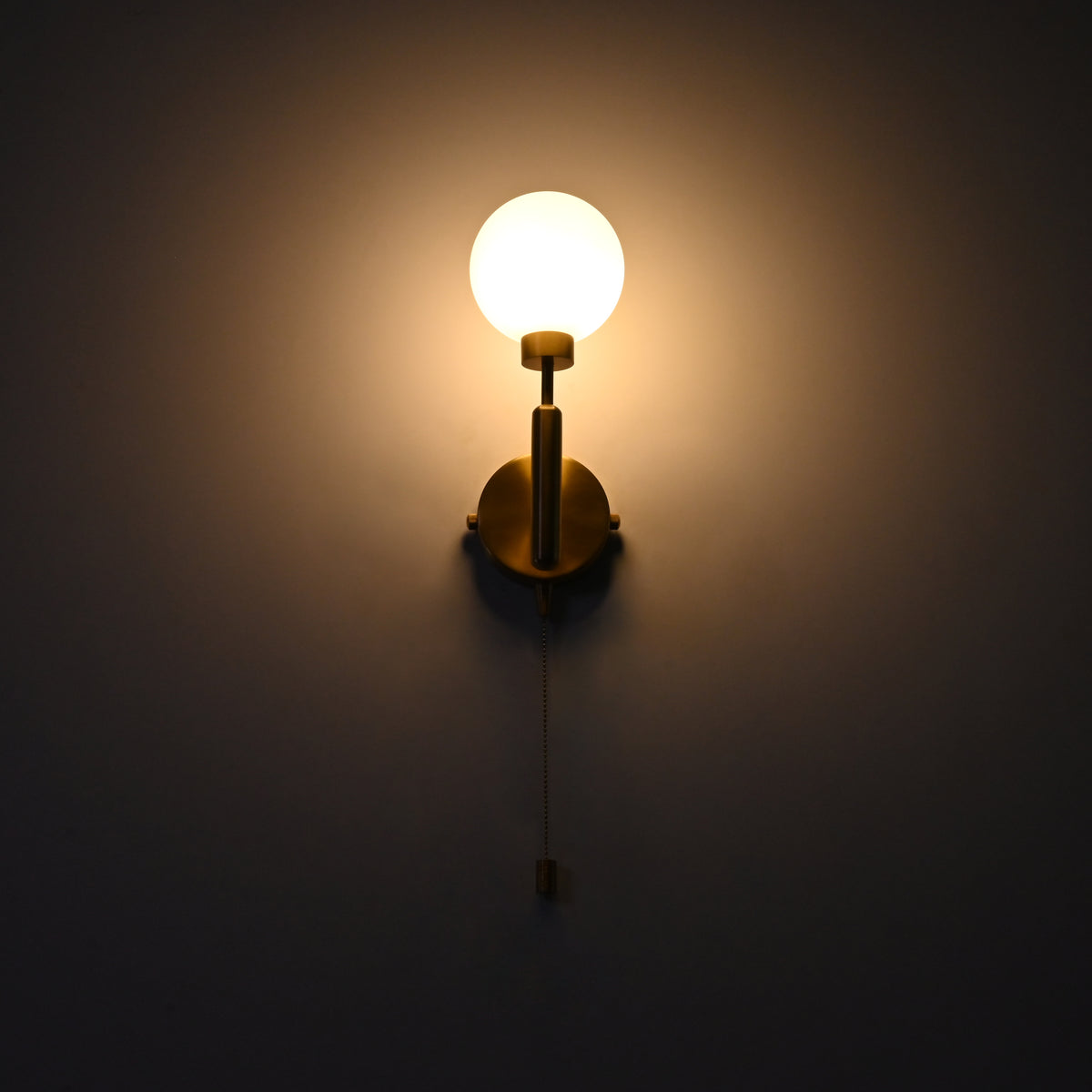 Raw brass wall sconce with a round frosted glass globe illuminated warmly, mounted on a dark wall with a hanging pull chain.