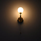 Raw brass wall sconce with a round frosted glass globe illuminated warmly, mounted on a dark wall with a hanging pull chain.